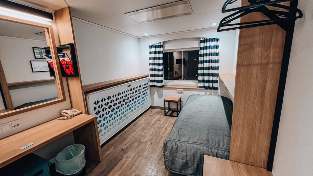The look of the Sija Serenade A class cabin. There is a window overlooking the sea with curtains. A single bed with a cover on it. hangers for your coats and a deck with a phone.