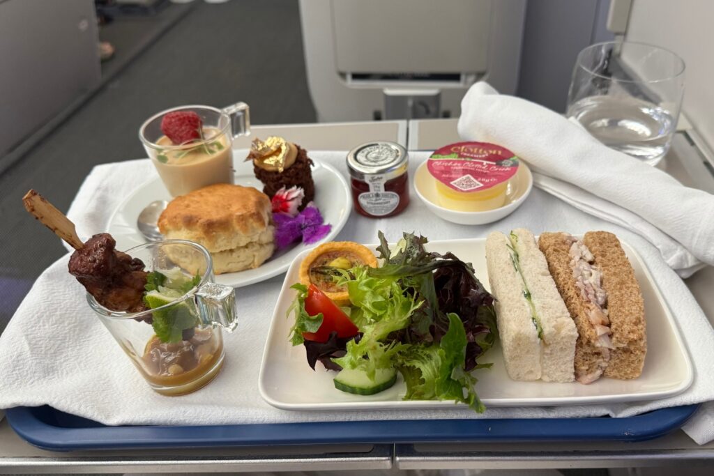 afternoon tea on an airplane table which includes sandwiches with salad, scone with jam and cream, a mousse dessert and some cake.